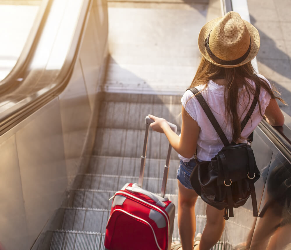 Frau mit Hut und Koffer fährt Rolltreppe runter