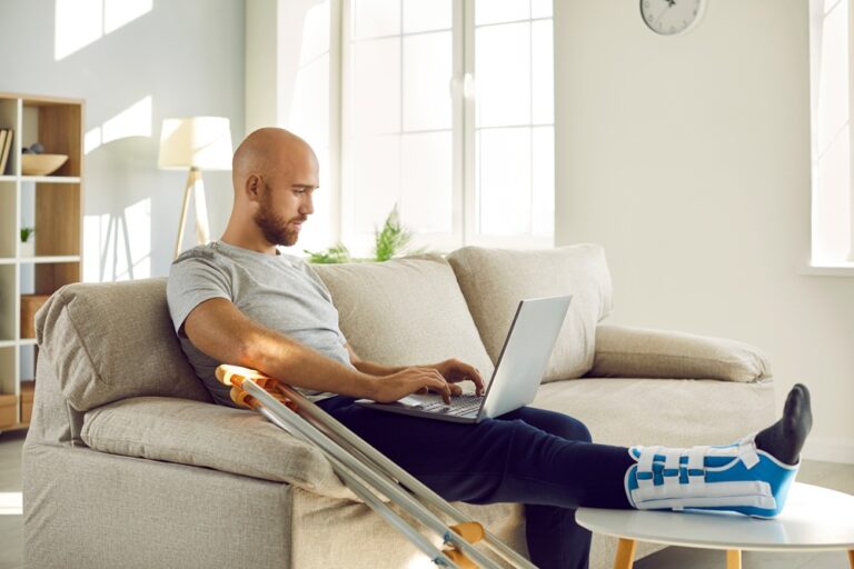 Mann mit gebrochenem Bein sitzt auf dem Sofa und arbeitet am Laptop